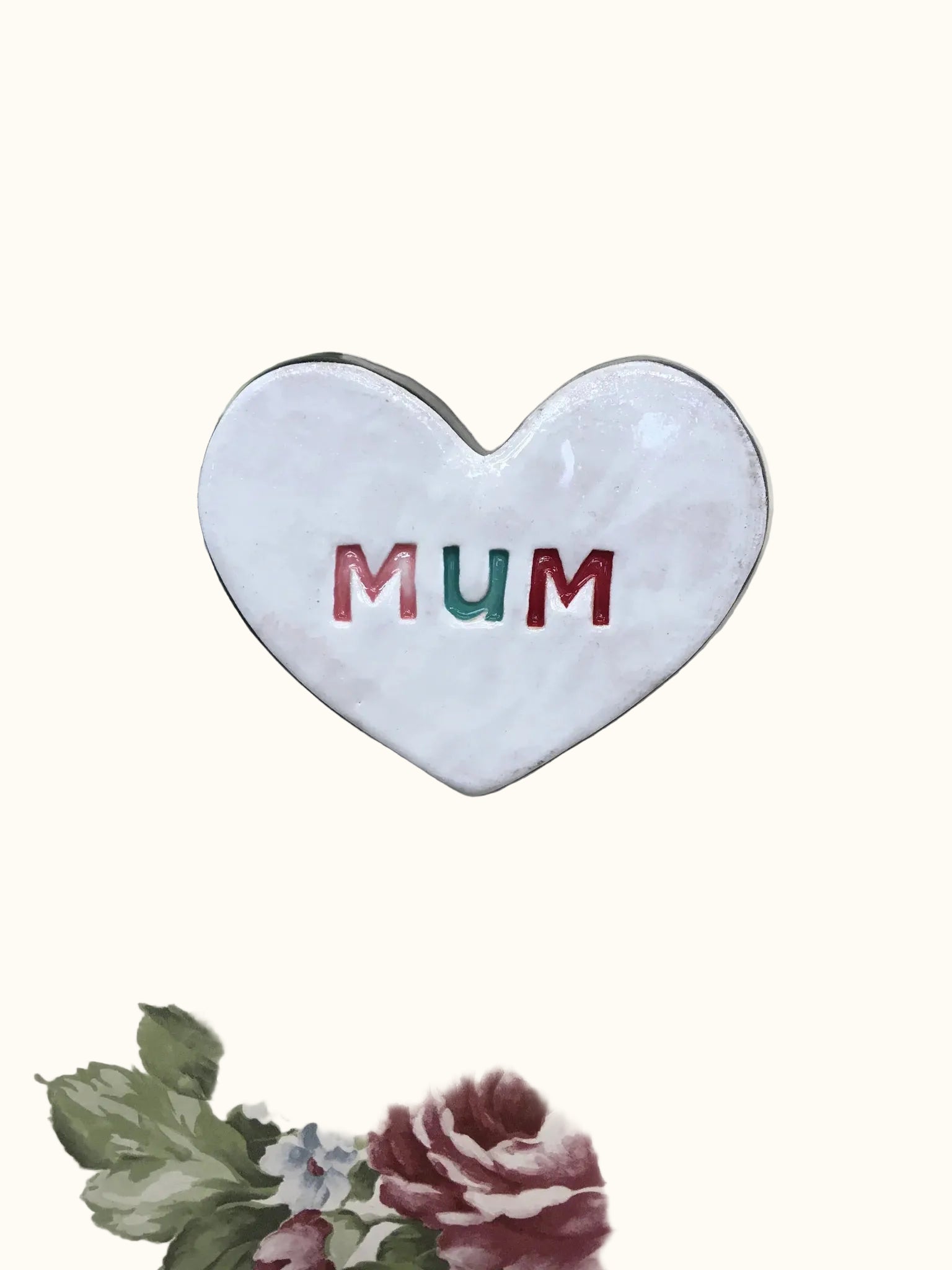 A heart-shaped ceramic ornament with the word 'MUM' stamped in the center, displayed on a floral background.